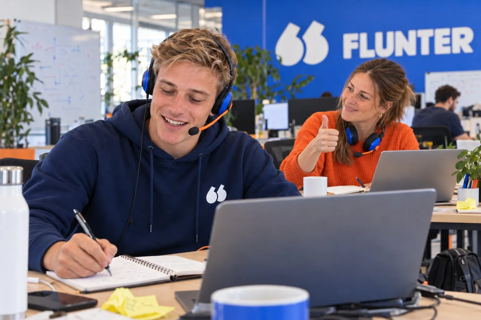 Un jeune SDR au casque note un rendez-vous pendant son appel, une collègue lui fait signe que c'est gagné
