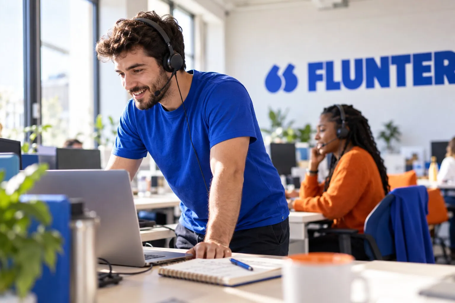 Un commercial debout au casque sur un plateau de prospection, une collègue en appel derrière lui